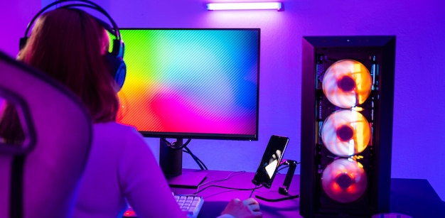 Asian professional gamer playing online video game on desktop computer pc have colorful neon led lights young woman in gaming headphones using computer for playing game at home back view