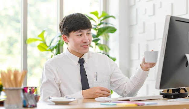 넥타이를 매고 공식적인 셔츠를 입은 아시아의 성공적인 남성 사업가 직원은 사무실 책상에 있는 컴퓨터 모니터에서 동료와 인사를 나누며 손으로 인사하는 커피 컵을 흔들고 있습니다.
