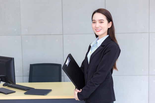 검은 양복을 입은 아시아 전문 여성은 손에 클립보드를 들고 자신감 넘치는 미소를 짓고 있다