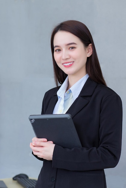 검은 양복을 입은 아시아 전문 직장 여성은 손에 태블릿을 들고 사무실에서 미소를 짓고 있다