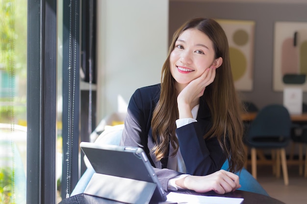 검은 양복을 입은 아시아 직업 여성이 테이블 위에서 행복하게 웃고 있는 태블릿 작업을 하고 있다