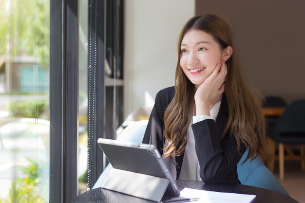 外を見て黒いスーツを着たアジアのプロの働く女性は、テーブルの上のタブレットに取り組んでいます