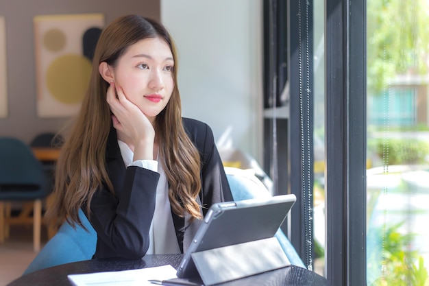 外を見て黒いスーツを着たアジアのプロの働く女性は、テーブルの上のタブレットに取り組んでいます