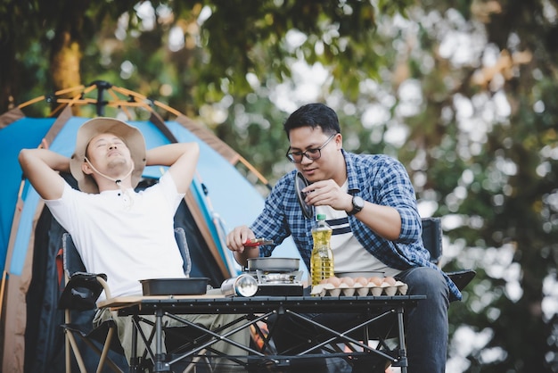 Asian relaxed man using smartphone and waiting for his friend cooking with tent background in camping Cooking set front ground Outdoor cooking traveling camping lifestyle concept