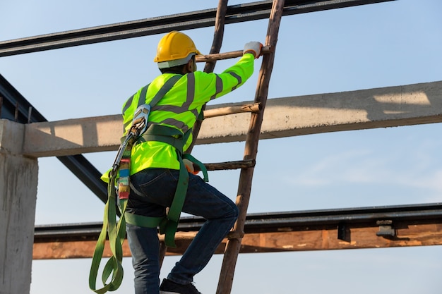 L'operaio edile asiatico indossa l'attrezzatura per l'altezza di sicurezza salendo le scale per installare il telaio del tetto, dispositivo anticaduta per il lavoratore con ganci per imbracatura di sicurezza in cantiere.