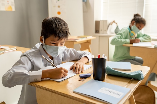 Asian schoolboy in protective mask writing in copybook