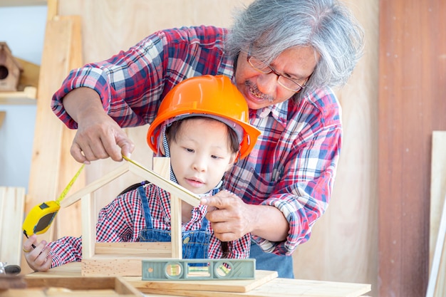 Asian senior carpenter man teaching asian little girl craftswoman measuring size of wooden house toy in carpentry shop.