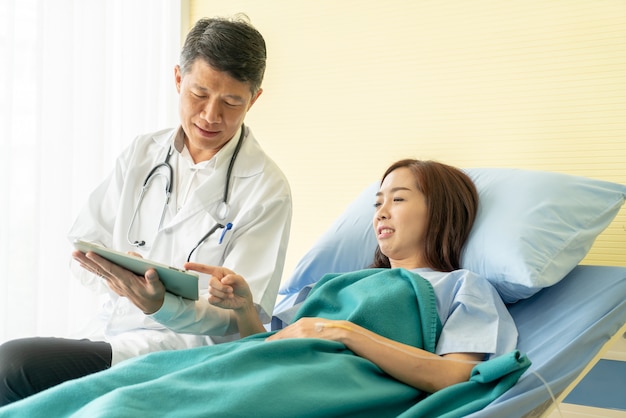 Asian senior doctor sitting on hospital bed and discussing with female patient