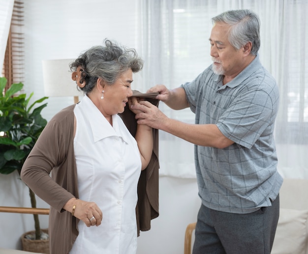 Asian senior elder man help elder woman wear a shirt at home.
