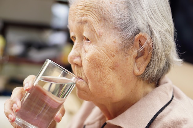 家のソファに座って水を飲むアジアのシニアまたは高齢の老婦人女性。ヘルスケア、愛、ケア、励まし、共感。