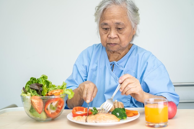 아시아 노인 또는 노인 여성 환자는 병원 침대에 앉아 배고픈 동안 야채 건강식으로 연어 스테이크 아침 식사를 먹고 있습니다.