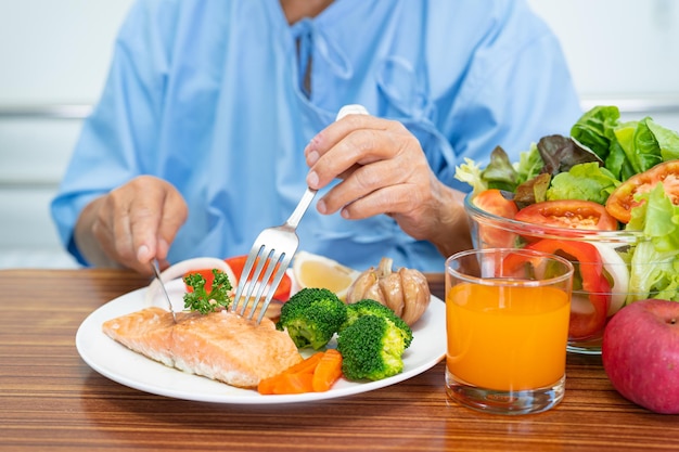 Paziente asiatico anziano o anziano che mangia bistecca di salmone colazione con cibo sano vegetale mentre è seduto e affamato sul letto in ospedale