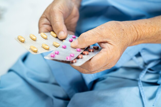 Asian senior or elderly old lady woman patient holding antibiotics capsule pills in blister packaging for treatment infection patient in hospital pharmacy drugstore concept