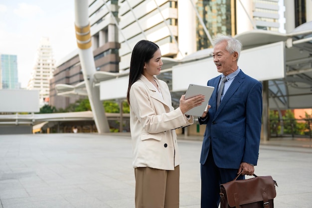 Asian senior mature middle aged businessman and young businesswoman having a discussion and using tablet in modern city