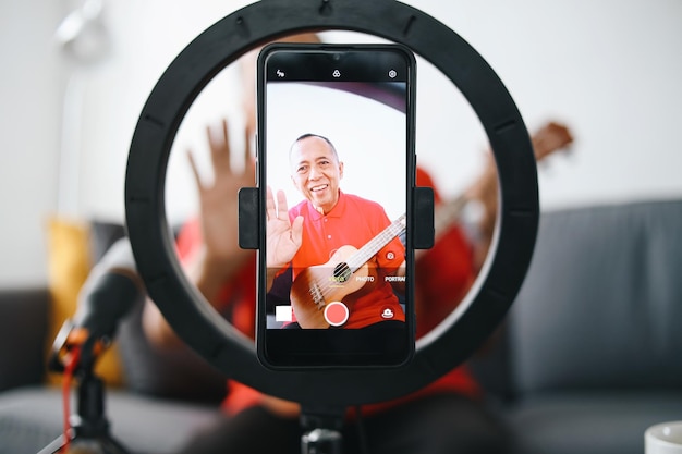 Photo asian senior old man playing guitar during podcast or live video broadcast at home