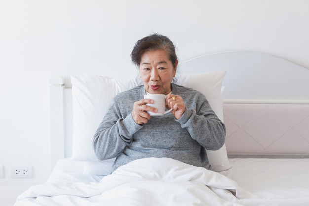 Asian senior old woman drinking coffee or tea on the bed