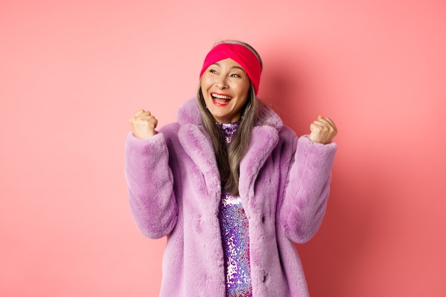 Asian senior woman celebrating, cheering and winning, making fist pump signs in triumph, achieve goal and smiling satisfied, standing in stylish faux fur coat, pink background.