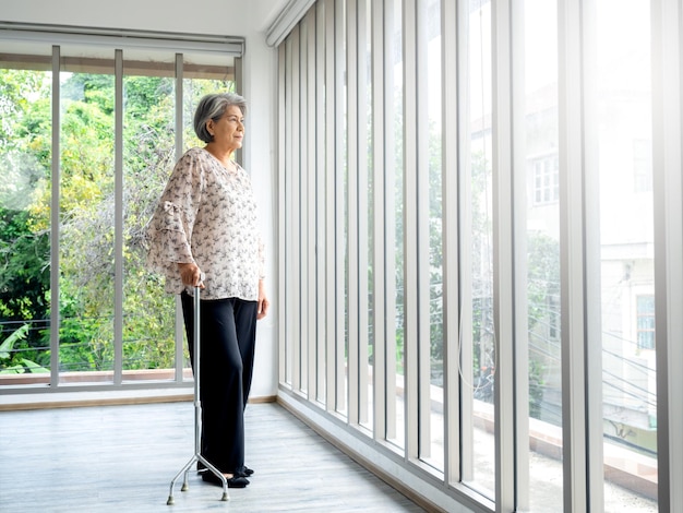 アジアの年配の女性の全身の白髪が杖を持って立ち、コピースペースのある室内のガラス窓の外を見ている 歩行杖を使った高齢の女性患者 強い健康医療のコンセプト
