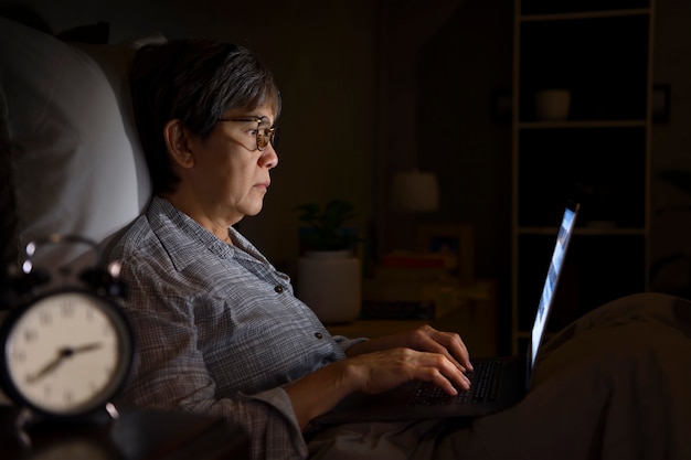 夜にベッドでノートパソコンを使用すると目が痛くて疲れているアジアの年配の女性