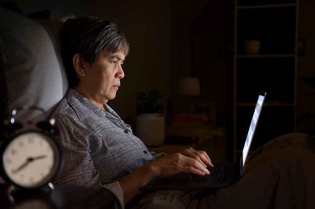 Donna maggiore asiatica che ha gli occhi irritati e stanchi quando si utilizza un laptop nel suo letto di notte