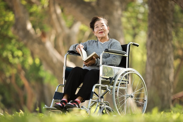 Азиатская старшая женщина сидя на кресло-коляске и читая книгу в улыбке сада сада и счастливом лице