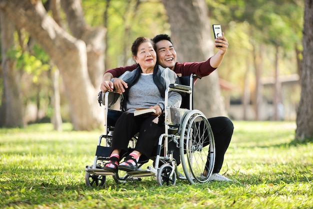 Donna senior asiatica che si siede sulla sedia a rotelle con suo fronte felice di sorriso del figlio sul parco verde