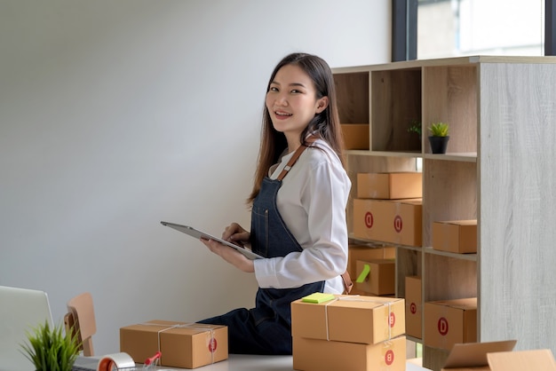 Asian small businesswoman holding tablet of products online before delivering them to customers with parcel box at home. Looking at camera.