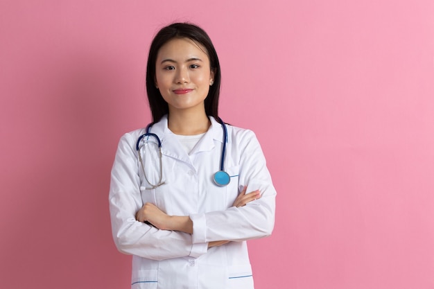 분홍색 배경 초상화에 청진기가 있는 흰색 의료 가운을 입은 아시아 웃는 의사 여자