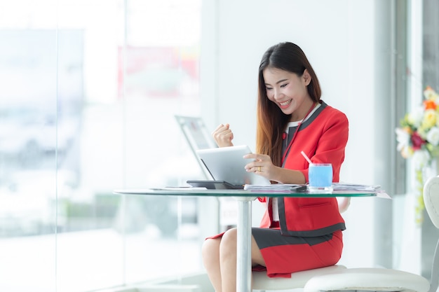 Asian smiling successful businesswoman in suit talking on phone using tablet sitting at of