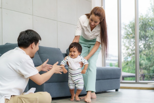 最初の一歩を踏み出したアジアの息子の赤ちゃんは彼の父に向かって歩きます。母親の助けを借りて歩くことを学び、家で優しく歩く方法を教える幸せな小さな赤ちゃん