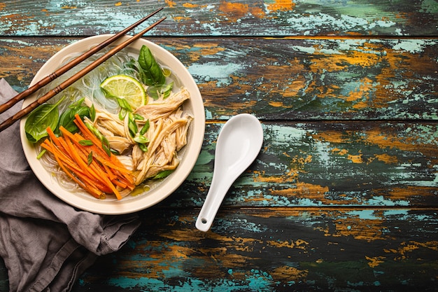 Zuppa asiatica con spaghetti di riso, pollo e verdure in ciotola di ceramica servita con cucchiaio e bacchette su fondo di legno rustico dall'alto con spazio per testo, cucina cinese o tailandese