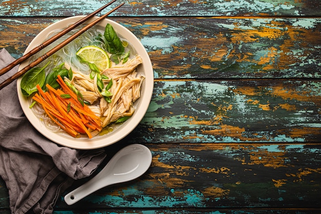 Zuppa asiatica con spaghetti di riso, pollo e verdure in ciotola di ceramica servita con cucchiaio e bacchette su fondo di legno rustico dall'alto con spazio per testo, cucina cinese o tailandese