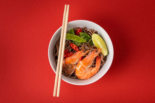 Asian stir fry soba noodles with shrimps, vegetables, green peas, red pepper in white bowl with wooden chopsticks, from above. Asian noodles on red background, close up, top view