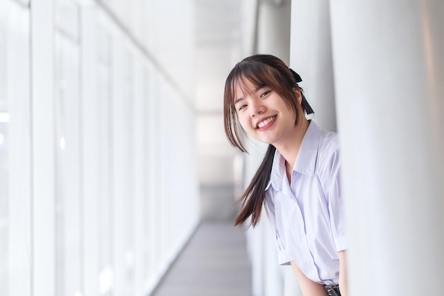 Studentessa asiatica in uniforme scolastica si alza e sorride felicemente con l'edificio scolastico.