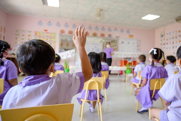 タイの学校の教室にいるアジアの学生幼稚園教師の質問に答えるために手を挙げている学生子供たちは好きで、学び、勉強を楽しんでいます