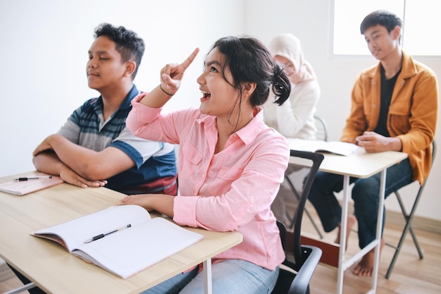 사진 아시아 학생들이 앉아서 손가락을 높이 가리키며 교실의 테이블에서 좋은 아이디어를 가지고 있습니다.