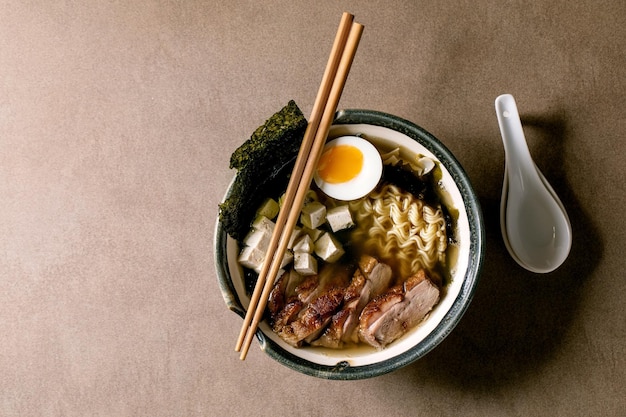 Asian style soup ramen with noodles grilled duck breast tofu seaweed nori chips and boiled egg in ceramic bowl with chopsticks and spoon on brown texture background Flat lay copy space