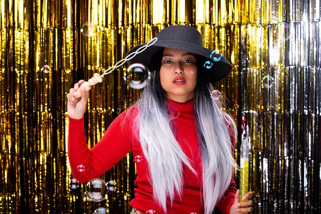 Asian Tan Skin girl wear dark Hat white hair red dress blows bubbles in Air, smiles and feel happy over golden foil curtain party with fun, studio lighting gold backgrounds copy space