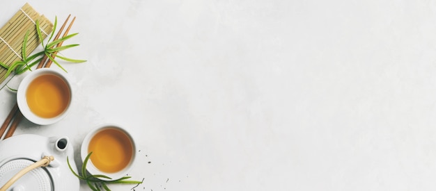 Asian tea concept, two white cups of tea, teapot, Tea set, chopsticks, bamboo mat surrounded with dry green tea on white background