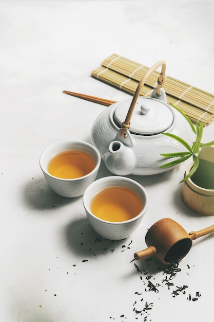 Asian tea concept, two white cups of tea, teapot, Tea set, chopsticks, bamboo mat surrounded with dry green tea