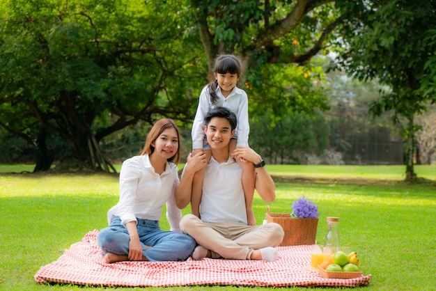 Asian teen family happy holiday picnic moment in the park with father, mother and daughter and smile to happy spend vacation time togerter in green garden with fruit and food.