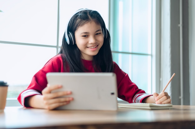 Studentessa asiatica adolescente indossa cuffie wireless studia online con riunione zoom, giovane donna felice impara la lingua ascolta lezione guarda webinar scrivi note guarda laptop istruzione a distanza.