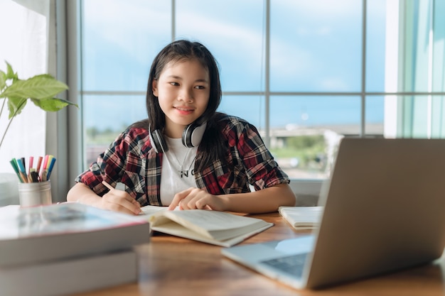 オンラインで言語を学ぶヘッドフォンを身に着けているアジアの十代の少女、ラップトップを使用して、画面を見て、自宅で学校のタスクを実行し、メモを書き、講義や音楽を聴き、遠隔教育