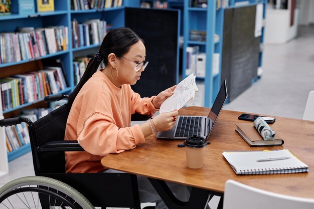 Asian teenage girl with disability studying in college library and using lap