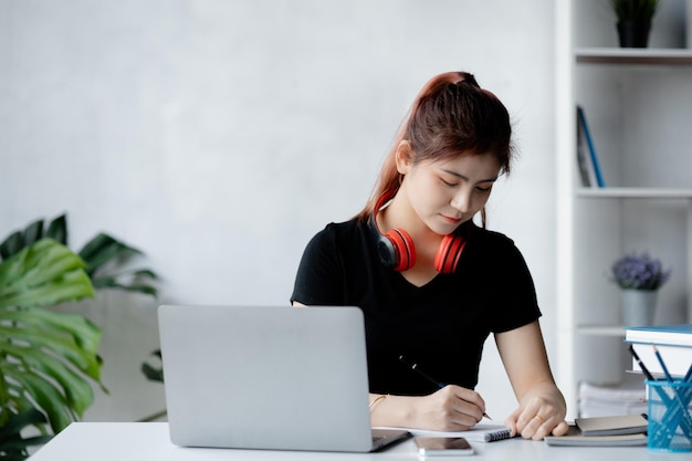 노트북을 들고 백인 사무실에 앉아 있는 아시아 10대 여성 그녀는 온라인 온라인 웹 교육 개념을 공부하는 가정 대학생에서 노트북으로 온라인 공부를 하는 학생입니다.
