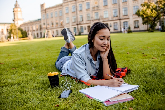 大学のキャンパスの近くの芝生に横になっているアジアの10代の女の子がノートにメモを書き込みます