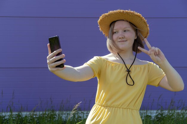 Foto la ragazza asiatica dell'adolescente in vestito e cappello gialli mostra il gesto di saluto mentre chatta video con gli amici