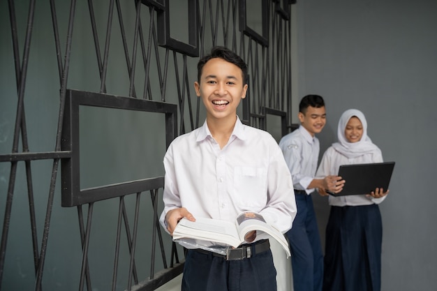 Un adolescente asiatico in uniforme scolastica sorride alla telecamera mentre porta un libro