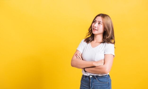 아시아계 태국인의 행복한 초상화 아름다운 귀여운 젊은 여성이 티셔츠를 입고 팔짱을 끼고 외진 곳을 바라보며 웃는 자신감, 노란색 배경에 스튜디오 촬영, 공간 복사