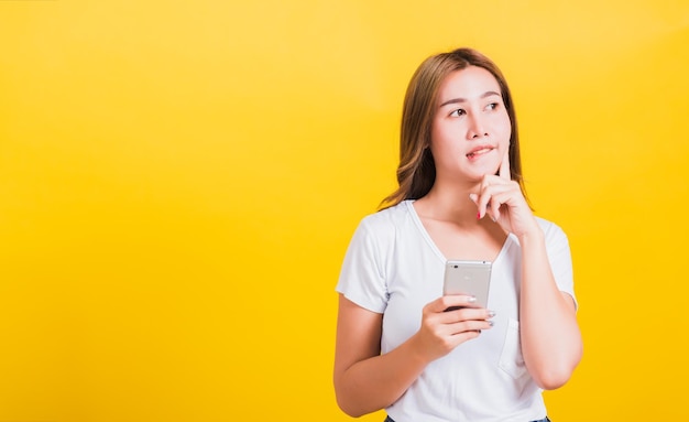 Asian Thai portrait happy beautiful cute young woman stand wear t-shirt her using holding smart mobile phone and chin handle thinking looking to side, studio shot isolated yellow background copy space